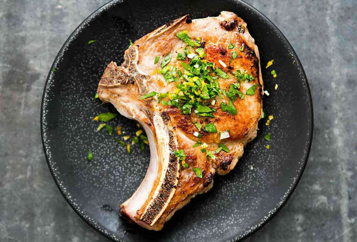 A cooked and seasoned pork chop served on a black plate, garnished with fresh chopped herbs.