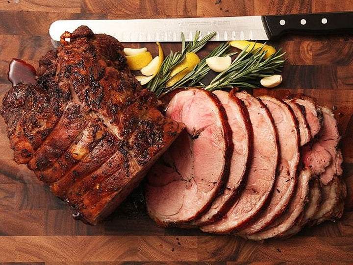 A succulent roasted beef joint, sliced on a wooden cutting board alongside fresh rosemary sprigs, lemon wedges, and garlic cloves, with a carving knife in the background.