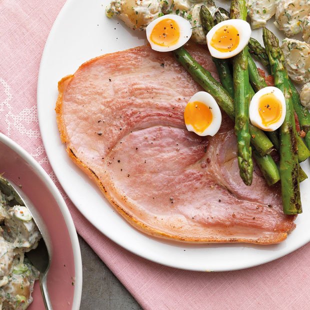 A cooked gammon steak served on a white plate with grilled asparagus, halved soft-boiled eggs, and a side of creamy potato salad.